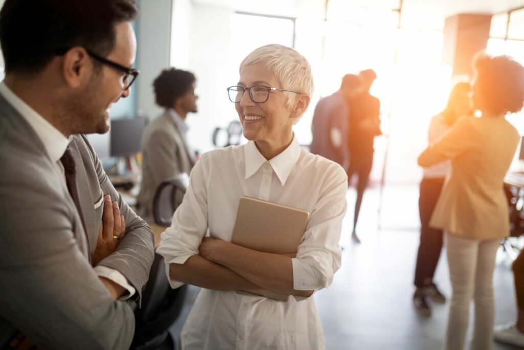 older white woman in a white business suite is smiling supportively at a white man in a business suit smiling back at her. Several blurred diverse professional figures in the background