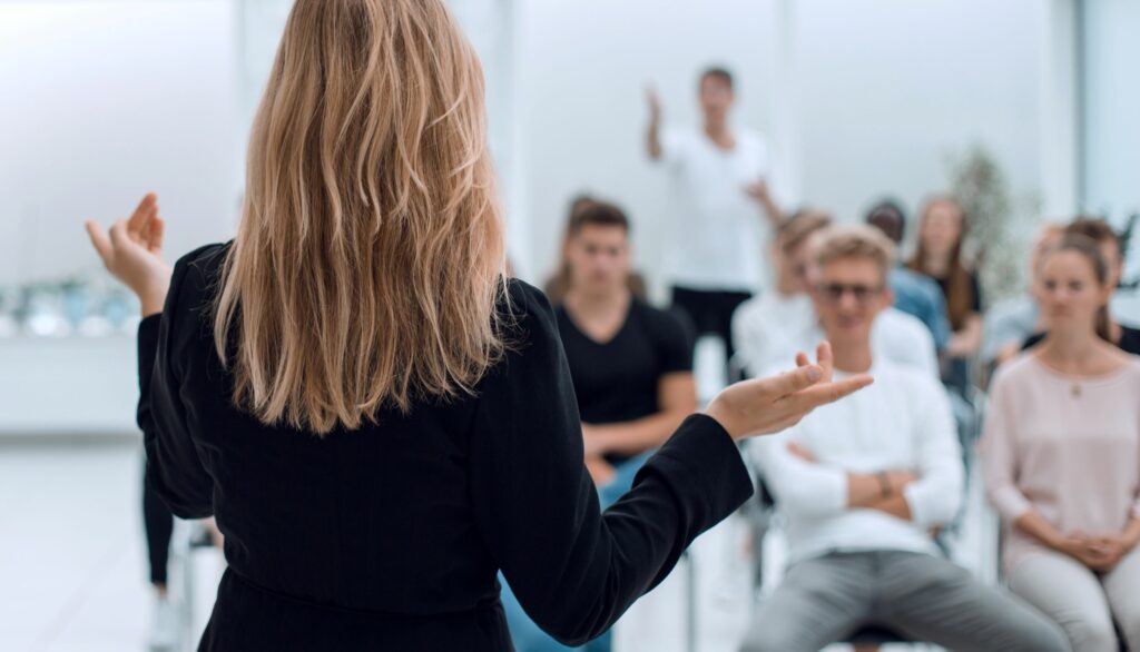 White woman with long light brown hair in a black business suit standing and facing a group of seated people