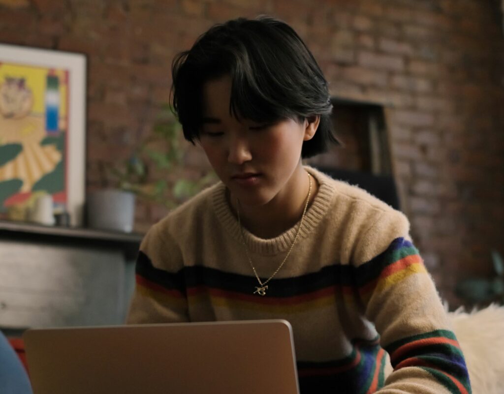 non-binary asian person wearing a sweater sits at home working on a laptop in the evening