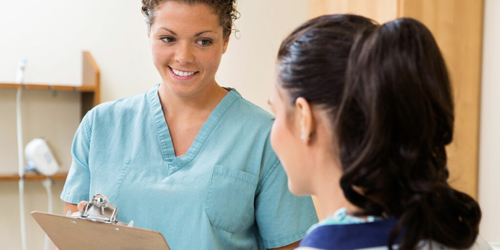 Young white woman in blue scrubs holding a clipboard speaking to another young White woman