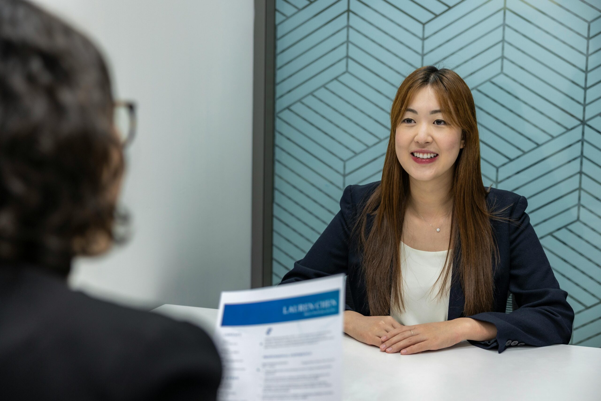 Young asian woman in professional dress sitting across a table from a woman, seen from the back, holding a document.
