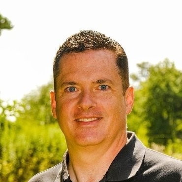 White man with short brown hair looking at the camera with a smile wearing a dark polo shirt against a natural background