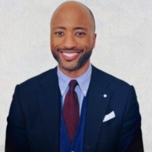 Black man, bald with a close beard wearing a dark blue suit and tie and a smile.