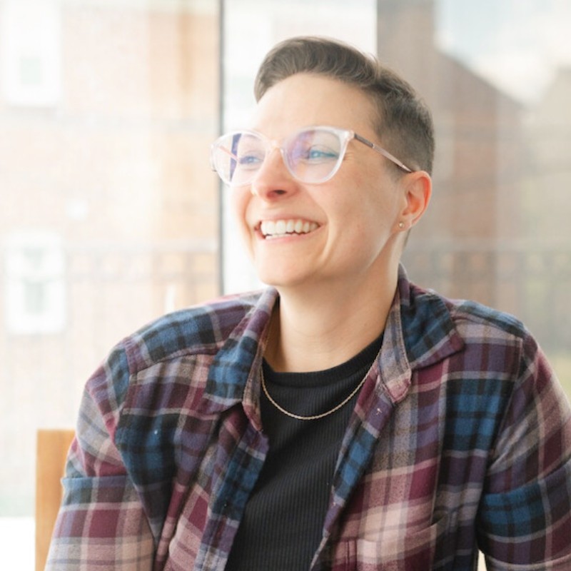 A smiling white woman with short brown hair wearing glasses, a plaid shirt and black t shirt.
