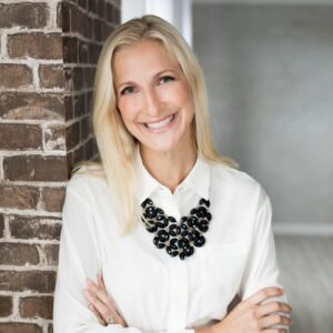 A white woman with blond hair smiling, wearing a white blouse with a chunky black necklace leaning against a brick wall.