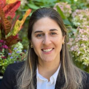 White woman with longer light brown hair facing the camera with a smile wearing a dark suit jacket and white shirt with a leafy background
