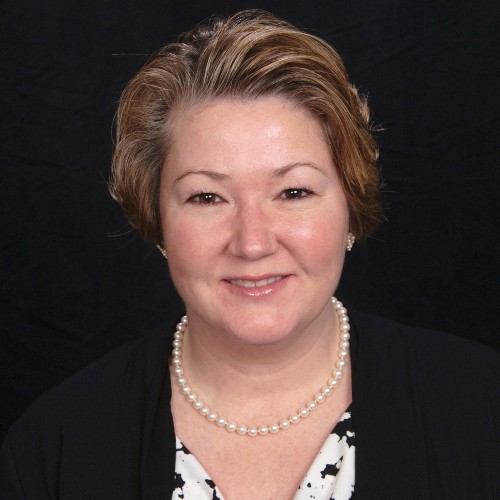 White woman with short blond hair looking into the camera wearing a black jacket and white patterned shirt on a black background