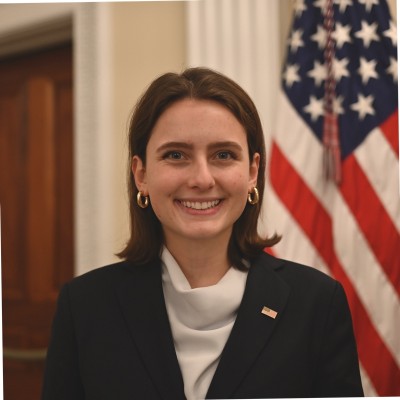 Young woman with medium length brown hair wearing a dark jacket and white turtleneck in front of an American flag.