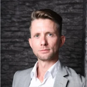 White man with brown hair brushed back in a wave wearing an open neck white shirt and gray jacket against a stone background