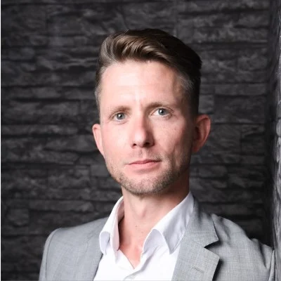 White man with brown hair brushed back in a wave wearing an open neck white shirt and gray jacket against a stone background