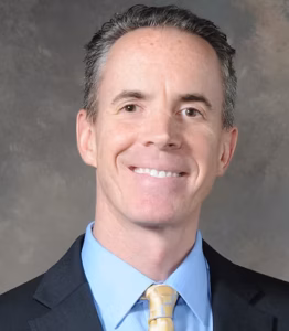 Middle aged white man with short brown hair smiling and wearing a suit jacket, shirt, and tie