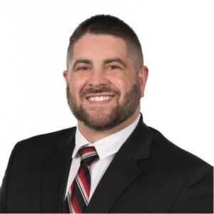 white adult male with short dark hair and close beard wearing a suite and tie