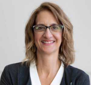 Smiling white woman wearing glasses and a white blouse and black jacket. She has dark blonde hair.