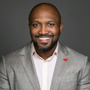 smiling black man with close cropped hair and short goatee and mustache wearing a gray suit and open-necked dress hirt