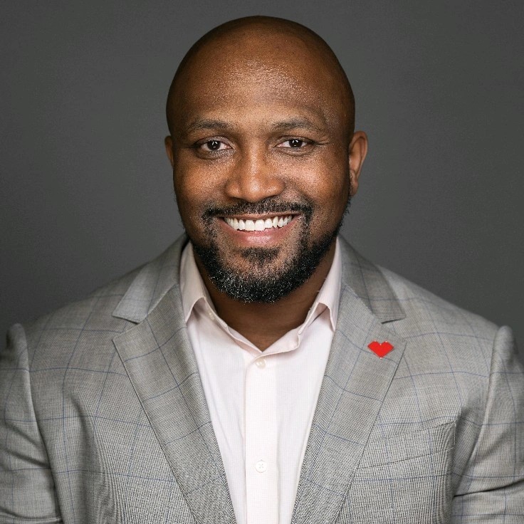 smiling black man with close cropped hair and short goatee and mustache wearing a gray suit and open-necked dress hirt