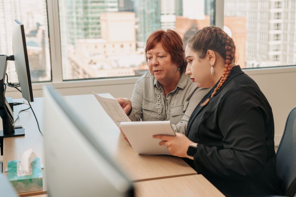 Older red-headed woman carefully reviewing a document next to a younger woman with a notebook listening.