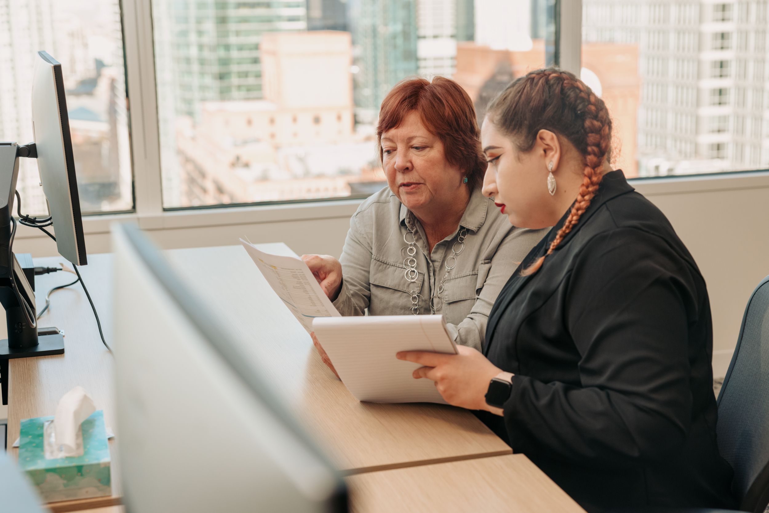 Older red-headed woman carefully reviewing a document next to a younger woman with a notebook listening.